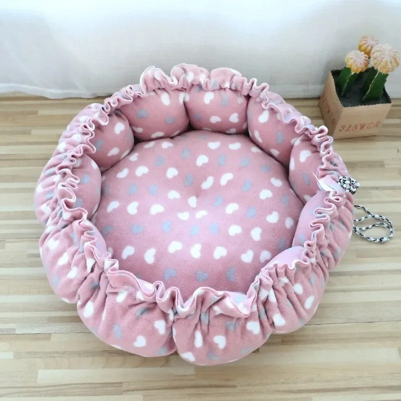 Un coussin chat en forme de nid avec un serrage tout le tour rose avec des motifs de cĆurs blancs et gris.