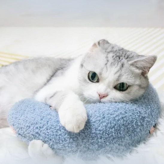 Un coussin pour chat en forme d'un oreiller croissant bleu avec deux fleurs roses et un chat gris allongé dessus et qui le tient avec ses deux pattes.