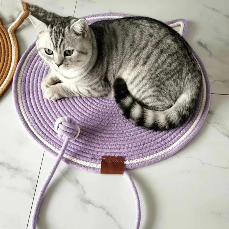 Un chat gris allongé sur un griffoir à chat en forme de tapis rond mauve avec un trait blanc tout le tour et une balle intégrée en corde de coton.