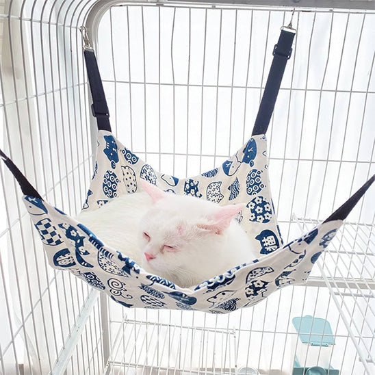 Hamac à chat balançoire blanc avec des motifs bleus accroché dans une cage avec un chat blanc endormis au-dessus.