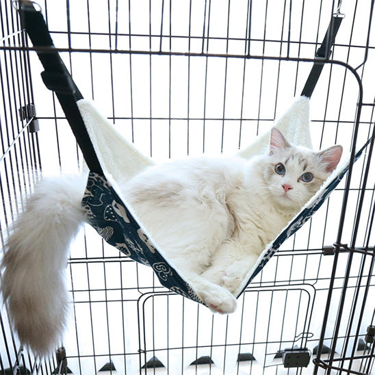 Hamac à chat balançoire bleu avec des motifs blancs accroché dans une cage avec un chat blanc allongé au-dessus.