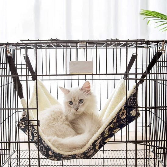 Hamac à chat balançoire blanc avec des motifs bleus accroché dans une cage avec un chat blanc assis au-dessus.