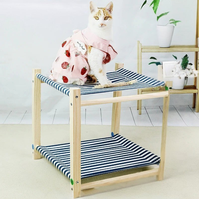 Un hamac à chat en forme de lit superposé en deux étages en bois et des sièges en toile à rayures bleue et blanc avec une chatte blanche portant une robe rose a motifs de fraises assise en haut.