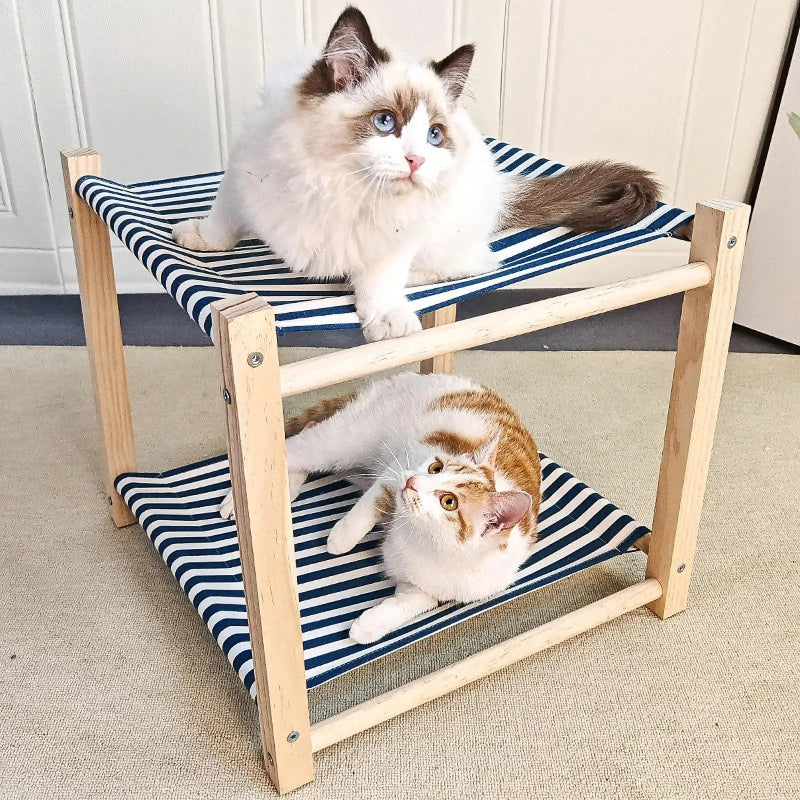 Un hamac à chat en forme de lit superposé en deux étages en bois et des sièges en toile à rayures bleue et blanc avec deux chats un ragdoll assis un en haut et l'autre blanc avec des taches rousses allongé en bas.