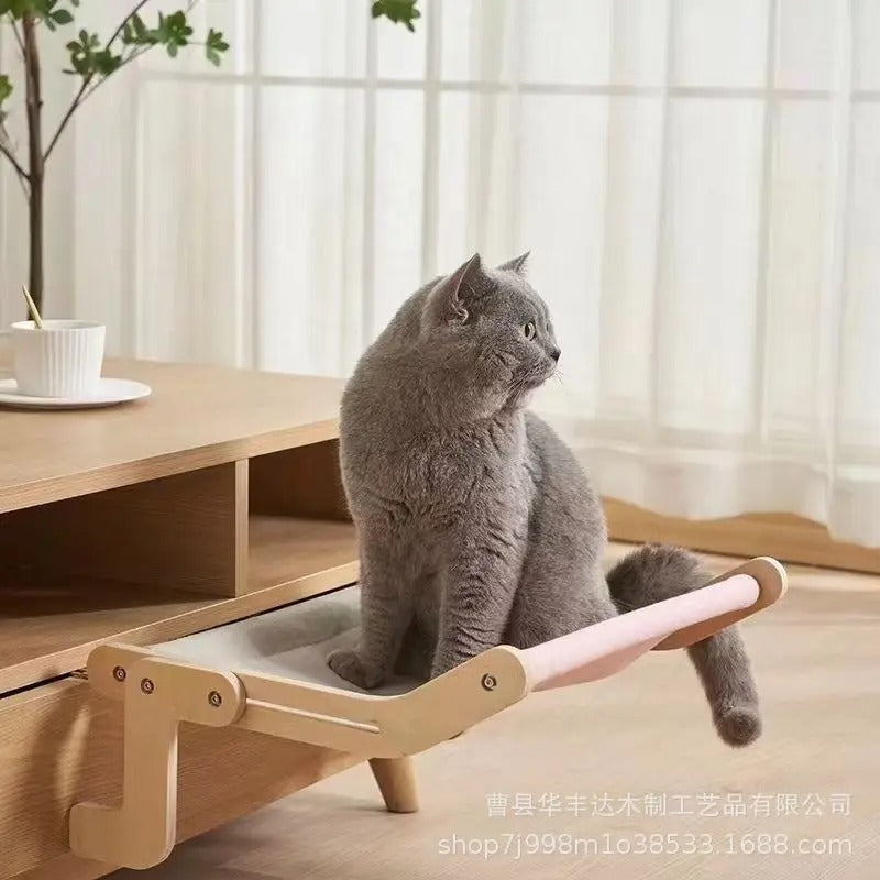 Un british shorthair assis sur un hamac chat gris et rose en bois accroché à un tiroir de table de salon et qui regarde ailleurs.