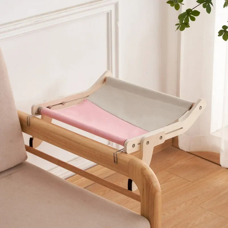Un hamac chat moitié gris moitié rose accroché à un accoudoir de fauteuil de salon en bois.
