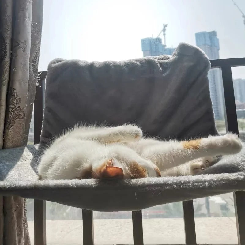 Un chat blanc qui dort sous le soleil sur un hamac pour chat gris placé sur le bord d'un balcon.