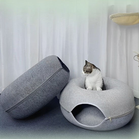 Lit pour chat en forme de donut gris clair et gris foncé avec un chat gris assis sur le lit gris clair.