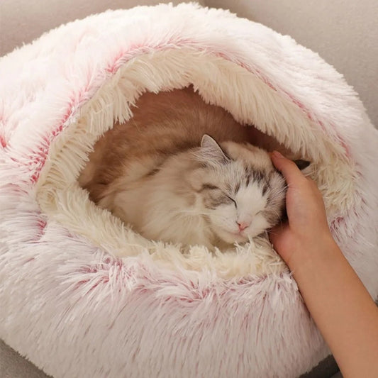 Lit pour chat en fore de cocon en peluche rose avec un chaton gris-blanc endormis dedans et une main qui le caresse.
