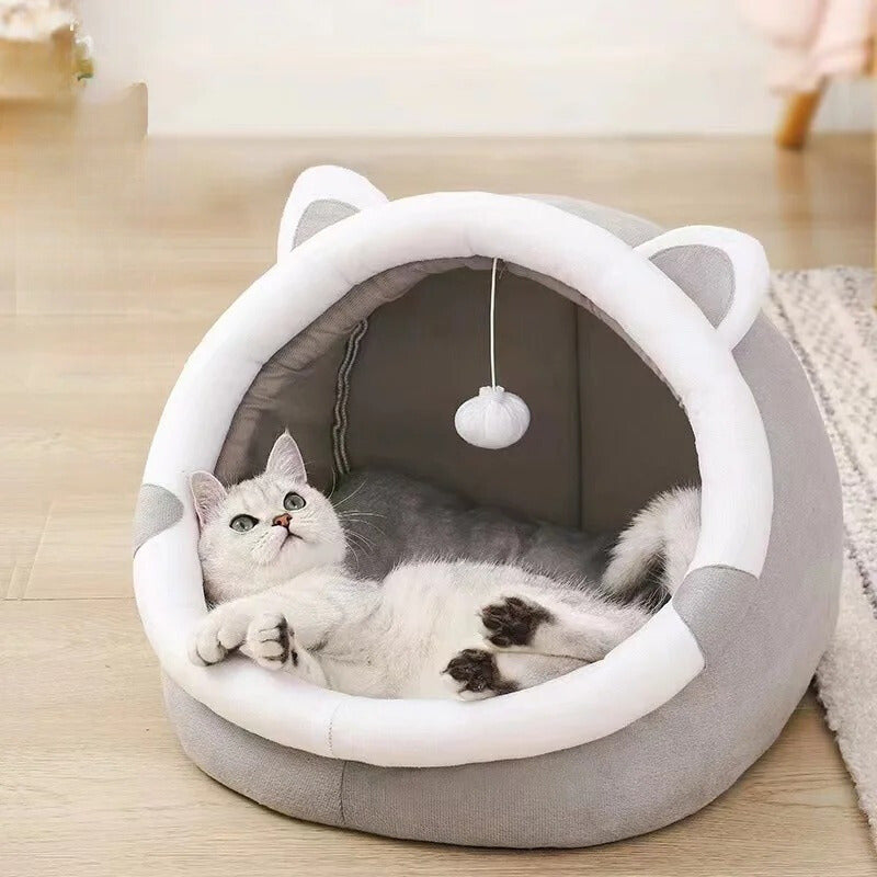 Niche pour chat grise en forme d'ourson avec des oreilles et une balle suspendue en haut et un chat blanc allongé dedans.