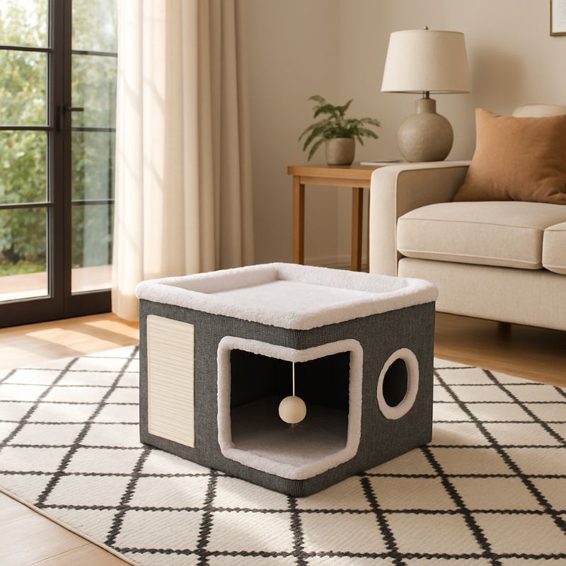 Niche pour chaton en forme de cube grise et blanche avec deux ouvertures, une zone de grattage et boule suspendue placée sur un tapis noir et blanc.