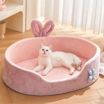 Un panier chat ovale rose avec deux oreilles de lapin et une fleur bleue sur le cÎté et une petite ouverture en avant et un chat blanc allongé dedans.