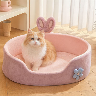Un panier chat ovale rose avec deux oreilles de lapin et une fleur bleue sur le cÎté et une petite ouverture en avant et un chat assis dedans.