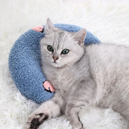 Un coussin pour chat en forme d'un oreiller croissant bleu avec deux fleurs roses et un chat gris allongé dessus.