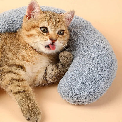 Un coussin pour chat en forme d'un oreiller croissant bleu avec deux fleurs roses et un chat marron allongé dessus en sortant sa langue.