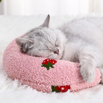 Un coussin pour chat en forme d'un oreiller croissant rose avec deux fraises tissées et un chat gris endormi dessus.