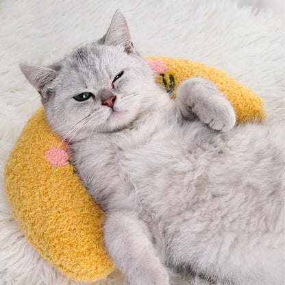 Un coussin pour chat en forme d'un oreiller croissant jaune avec deux fleurs roses et un chat gris allongé dessus.