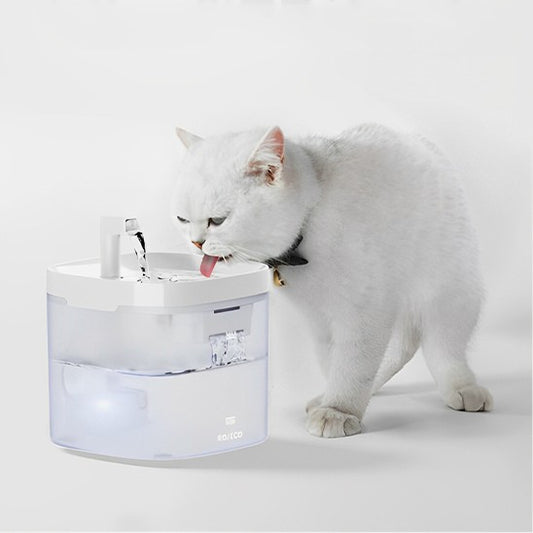 Une fontaine a eau chat automatique blanche et bien profonde en haut et transparente en bas avec un petit robinet et un chat blanc qui boit de cette fontaine.