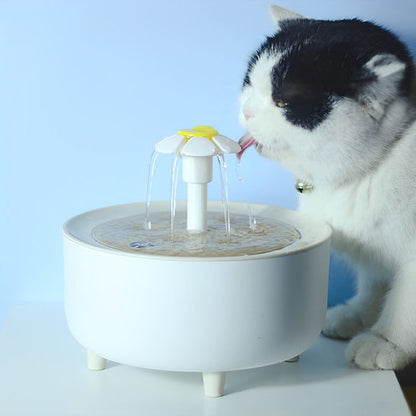 Une fontaine à eau pour chat ronde avec une tête en forme de fleur de marguerite qui fait sortir l'eau de plusieurs directions avec un chat blanc avec un peu de noir sur le visage qui boit de cette fontaine.