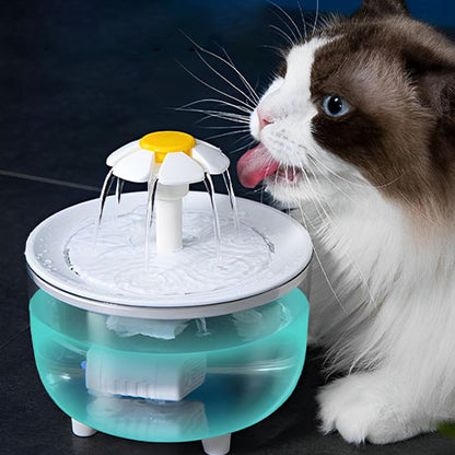 Une fontaine à eau pour chat ronde avec une tête en forme de fleur de marguerite qui fait sortir l'eau de plusieurs directions avec un chat blanc avec un peu de marron sur le visage qui tend sa langue pour boire de cette fontaine.