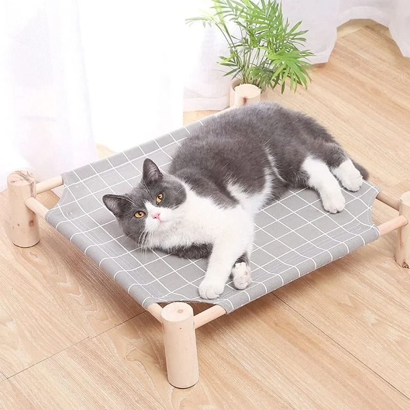 Un hamac à chat en forme carrée en bois surélevé du sol avec un tissu gris avec des carreaux blancs et un shorthair allongé dedans.