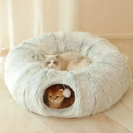 Lit pour chat en forme de tunnel fermé gris clair avec une balle suspendue et un chat roux assis dedans et un blanc assis dessus.