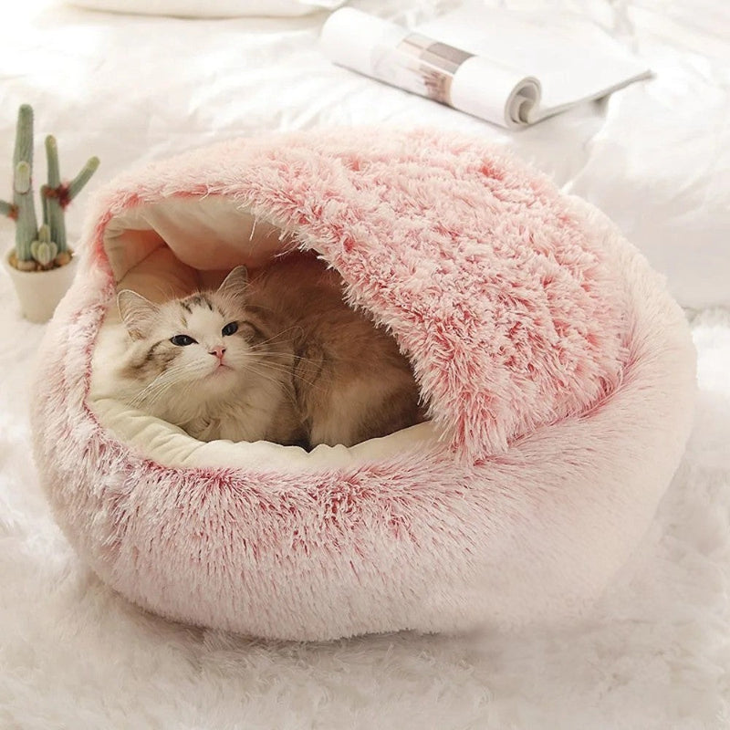 Lit pour chat en forme de petite grotte semi-fermée en peluche rose avec un chat blanc allongé dedans.