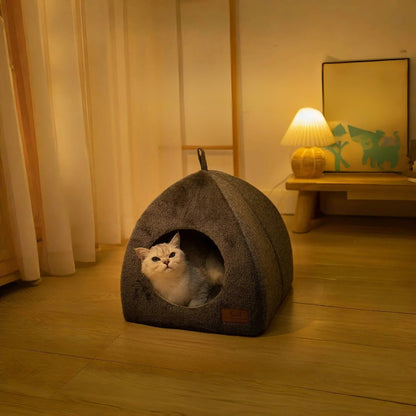 Maison pour chat en forme de petite niche grise avec un chat blanc assis dedans mise dans une pièce avec une lumière tamisée.